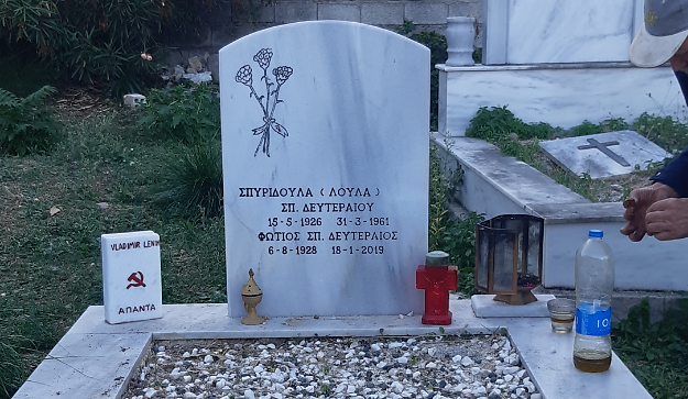 A tomb with a standing slab without a cross, bearing names and a bundle of three carnations. Next to it, exist a marble book model with an image of a hammer and sickle. 